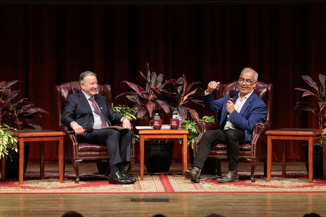 Two men sit in leather armchairs on a stage, with one speaking into a microphone while gesturing.