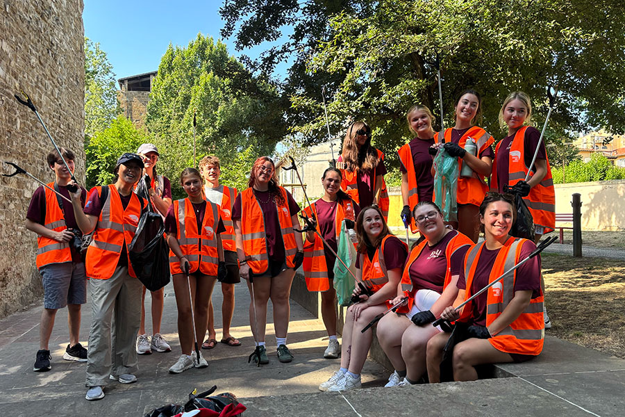 A group of students wearing orange safety vests poses in a shaded area in front of trees.