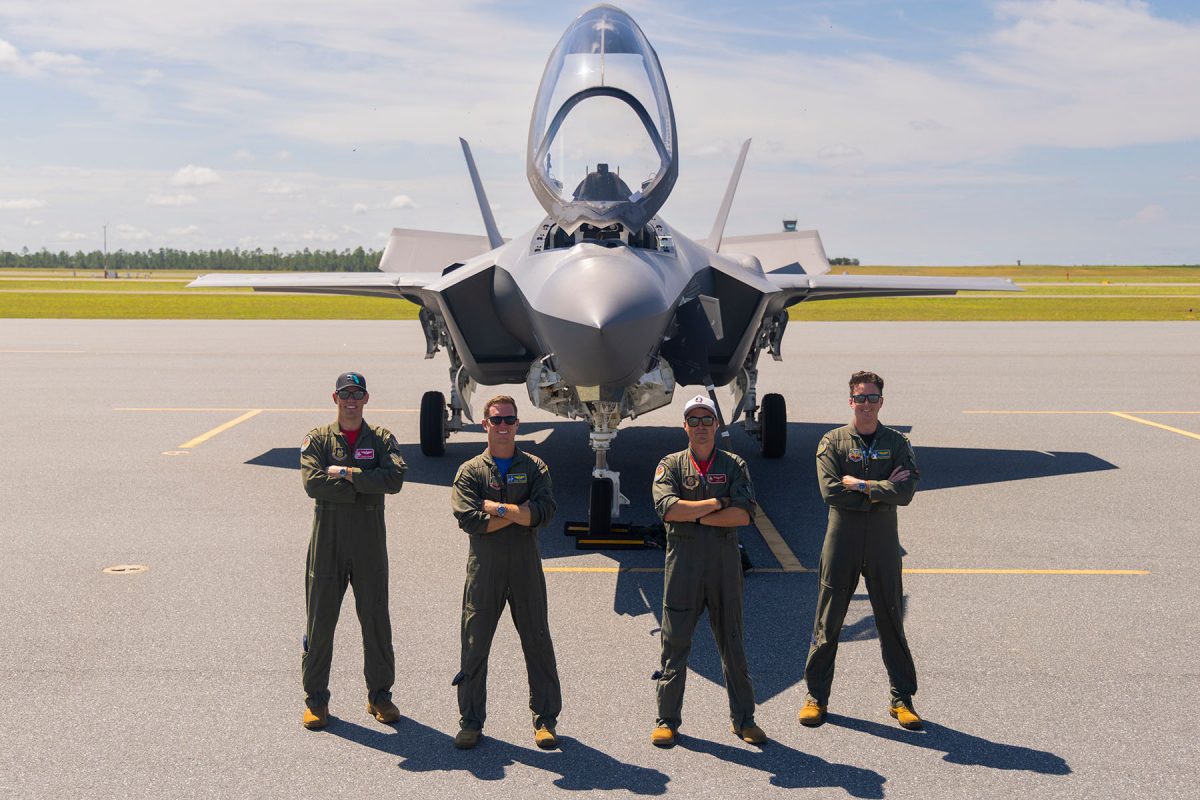 Four military pilots stand in flight suits with crossed arms in front of a fighter jet on a tarmac.