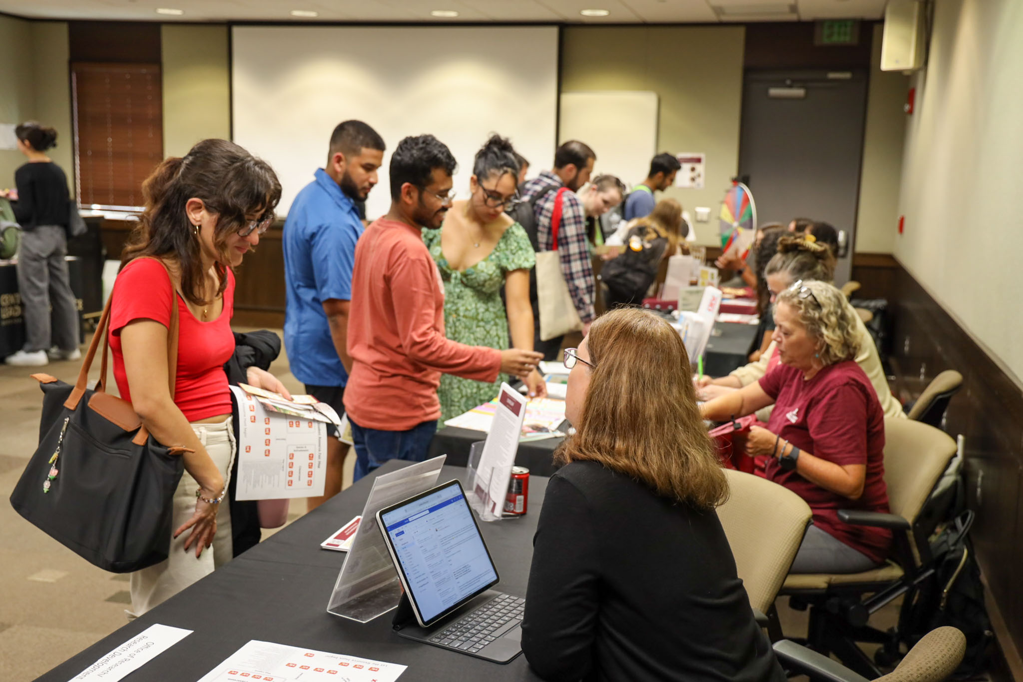 Students browse informational tables and speak with representatives at an indoor campus fair or orientation event.