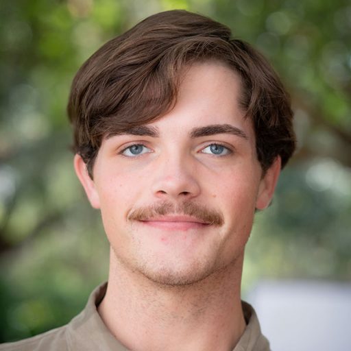 A man poses for a headshot with greenery out of focus behind him.
