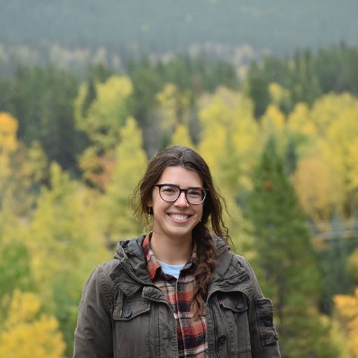 A woman stands with a forest behind her.