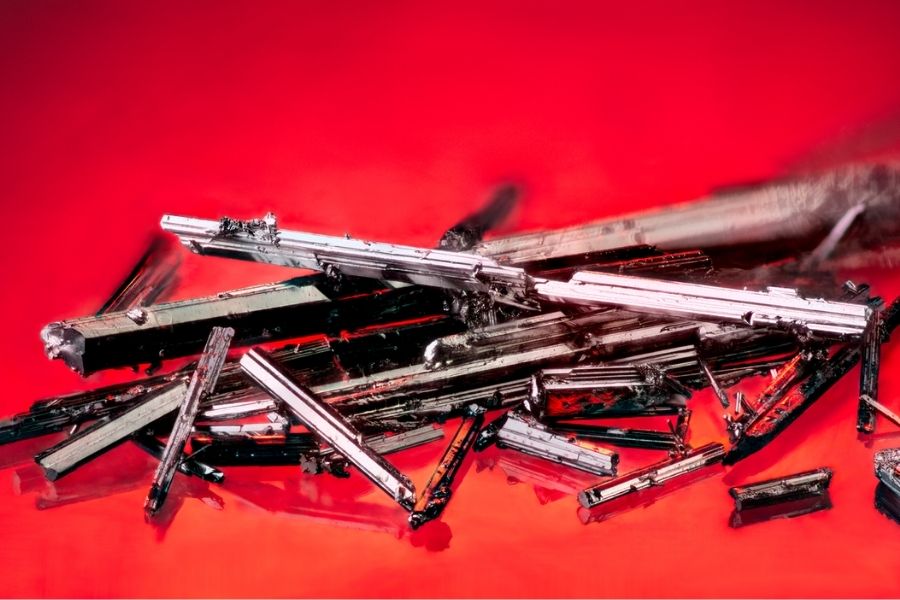 A close-up view of long, grey crystals in front of a red background.