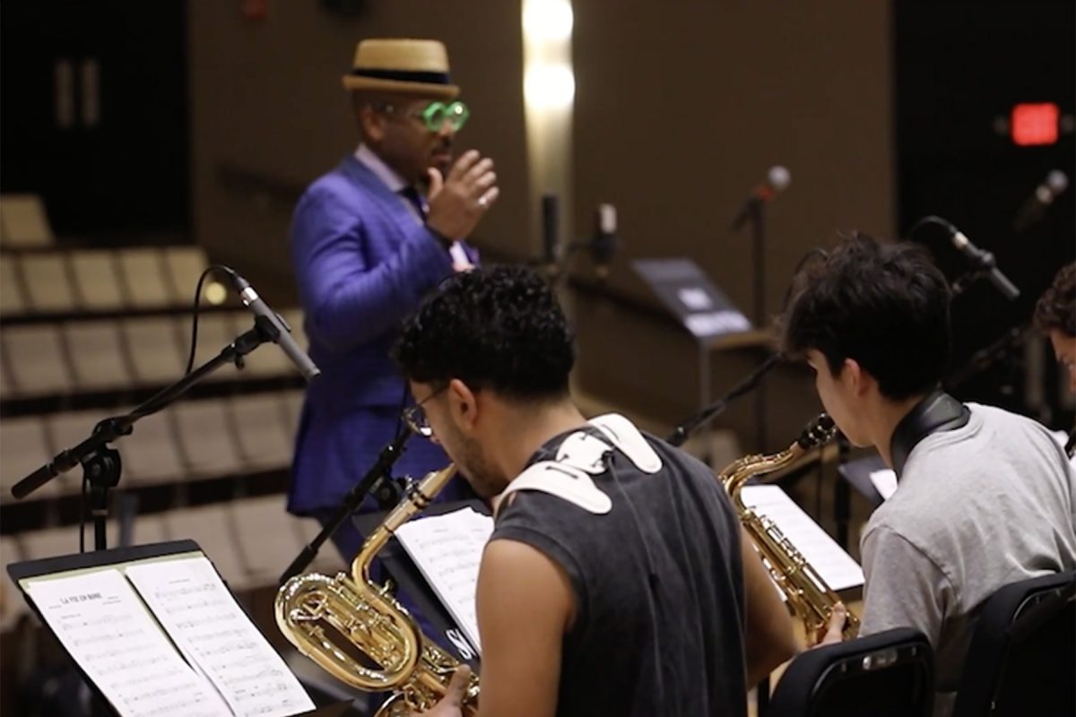 A man in a blue suit, hat, and bright green glasses directs a jazz ensemble while young musicians play saxophones with music sheets in front of them.