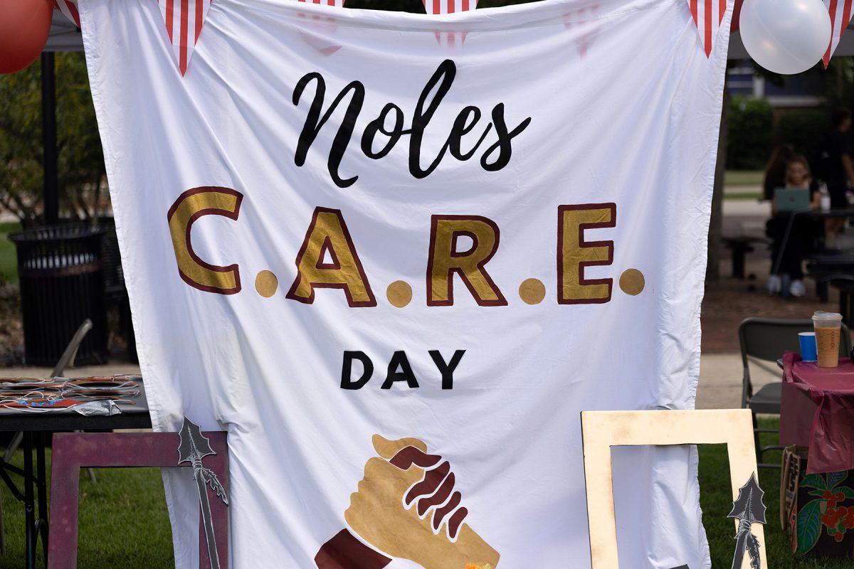 A white banner displaying the words "Noles C.A.R.E. DAY" above an illustration of holding hands hangs outdoors framed by decorative pennant bunting and balloons.
