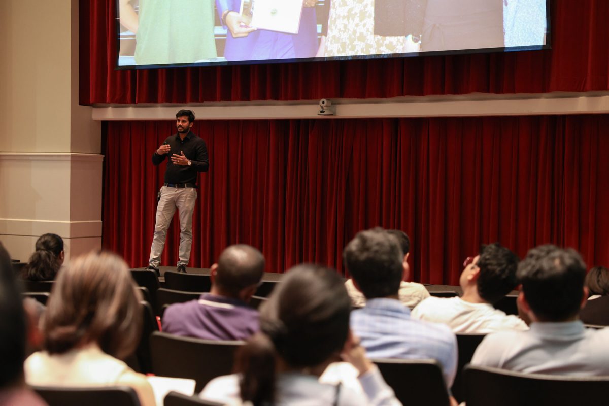 A man speaks on a stage in front of a red curtain and a large screen while the audience watches from auditorium seating.