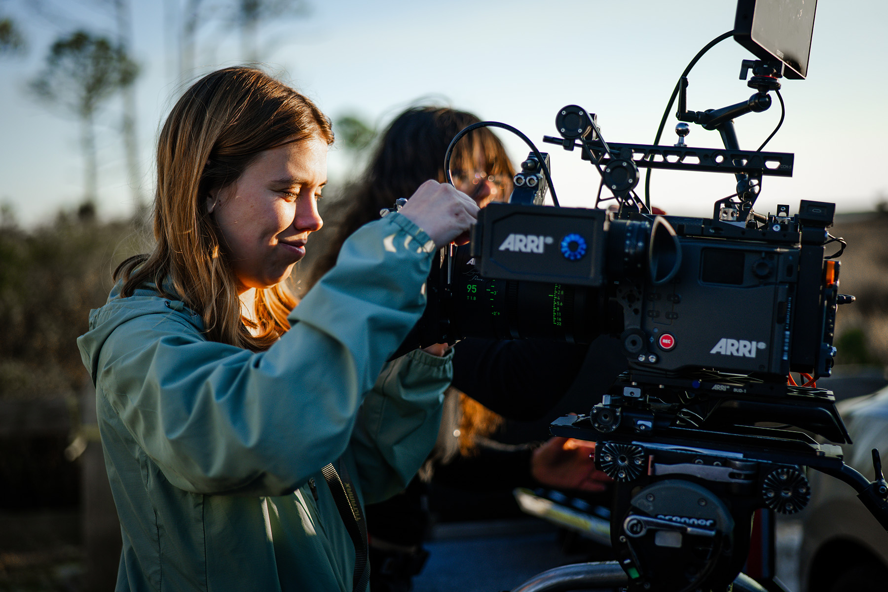 A student adjusts an ARRI camera on a rig outdoors while another person stands behind her on a film set.