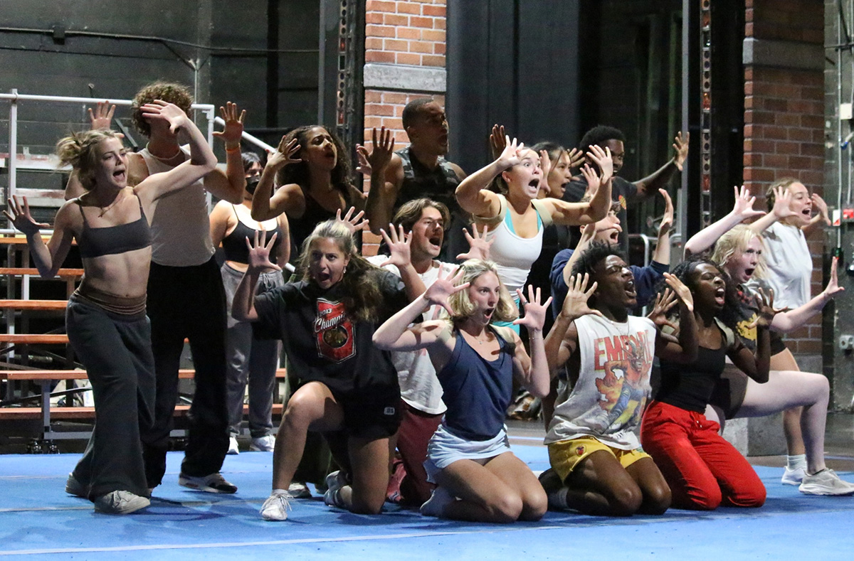 Student performers strike energetic poses with their hands raised and expressive faces during a musical rehearsal onstage.