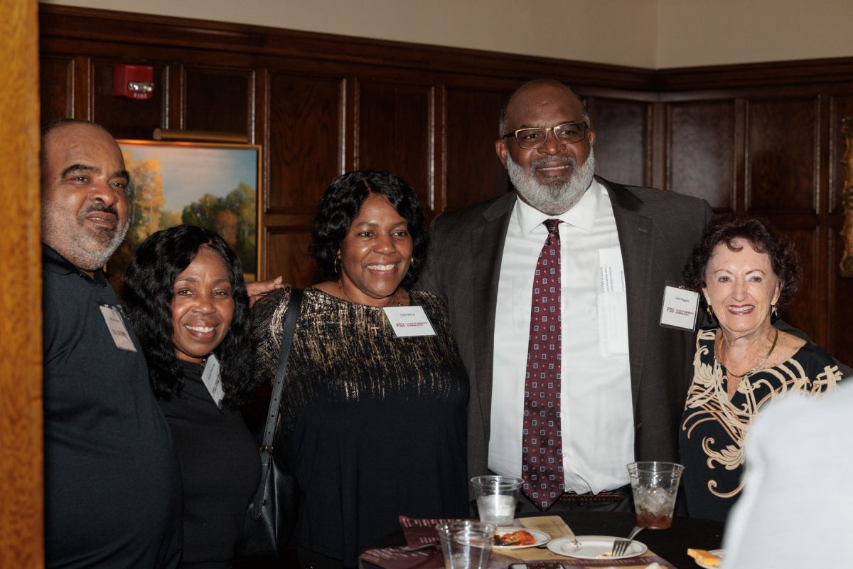 Odell Haggins, Jr. smiles while posing for a group photo with four other guests standing around a high-top table during a reception.