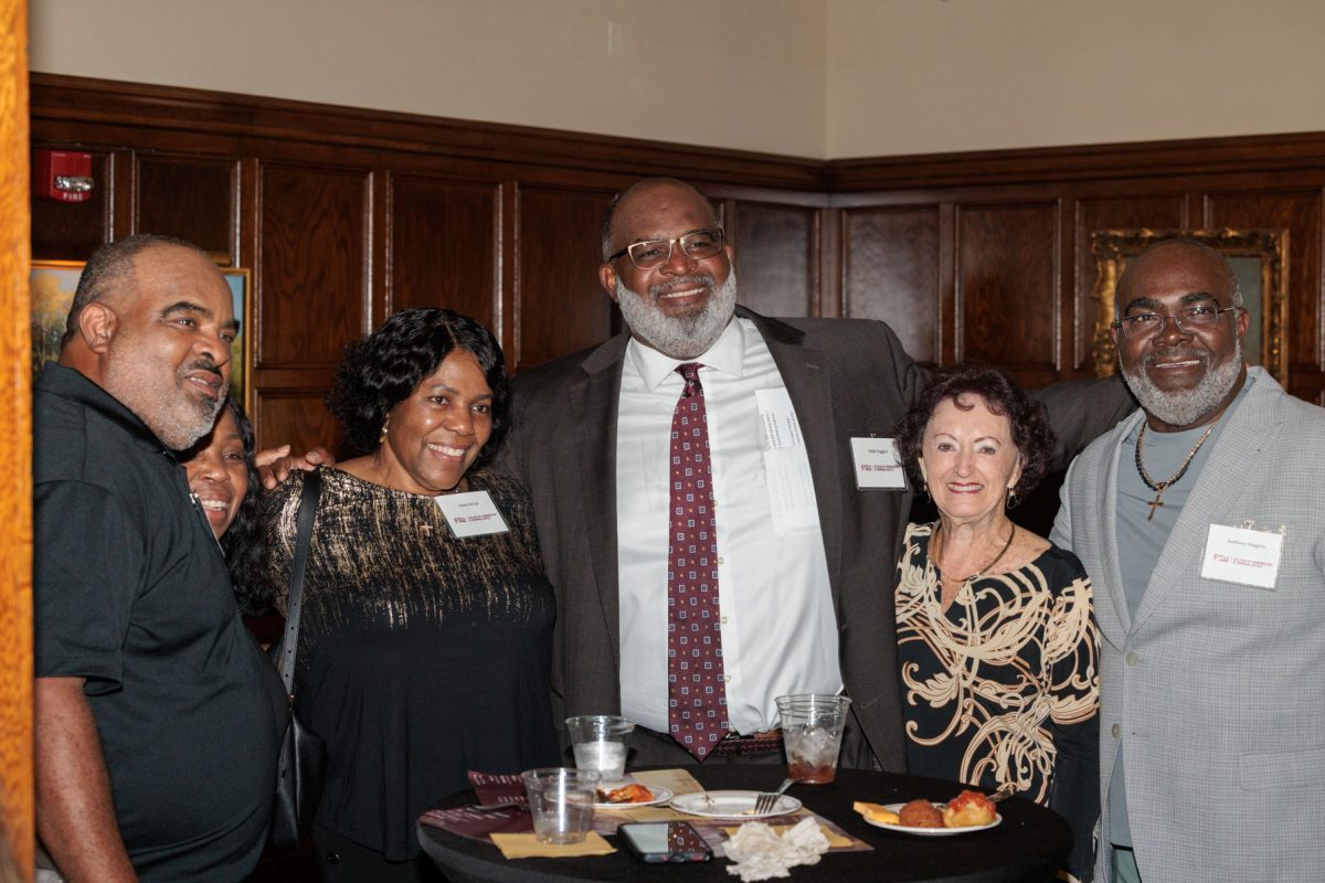 Odell Haggins, Jr. smiles with a group of five attendees standing around a small high-top table during a reception.