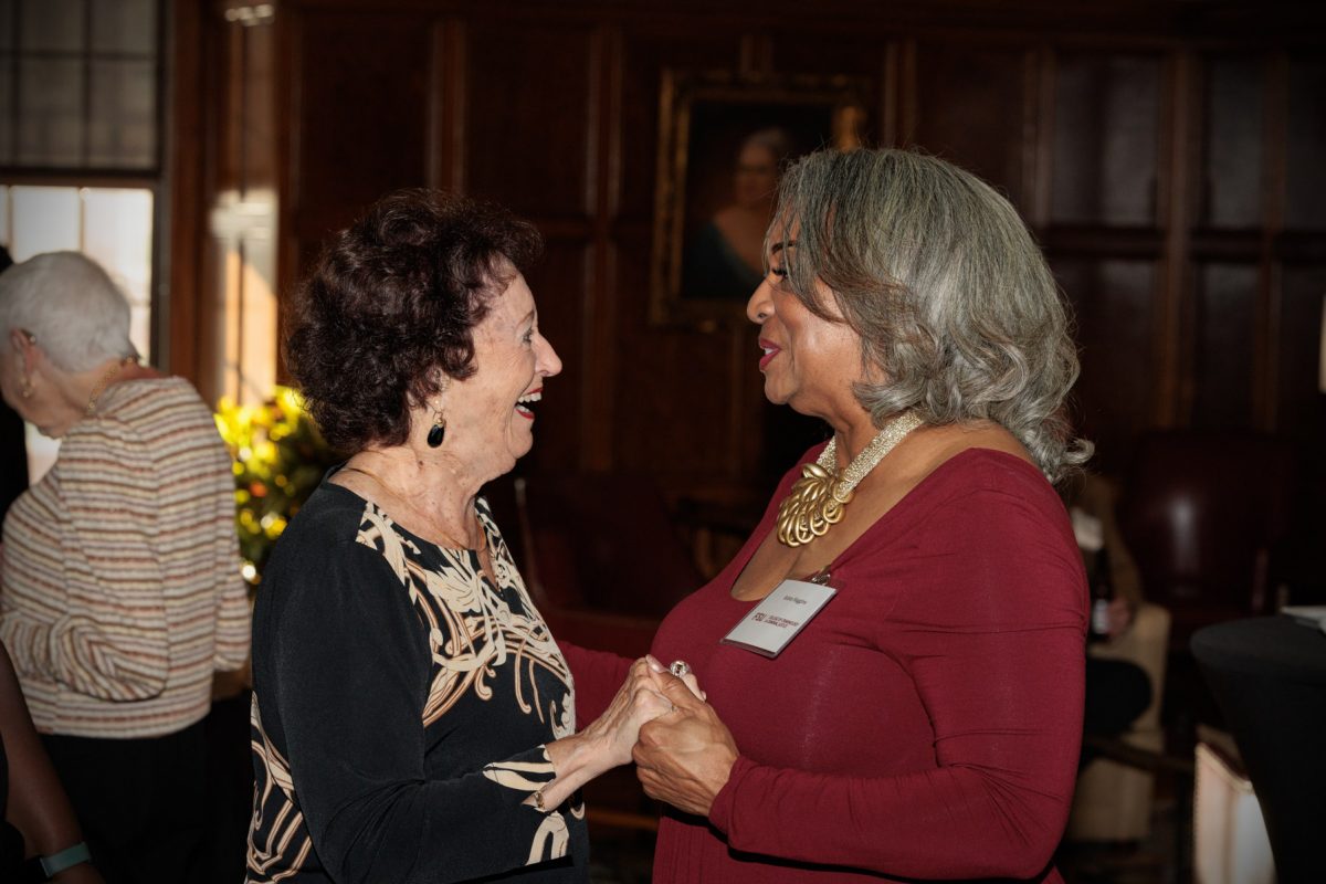 Two women smile and hold hands while conversing at a reception in a wood-paneled room.
