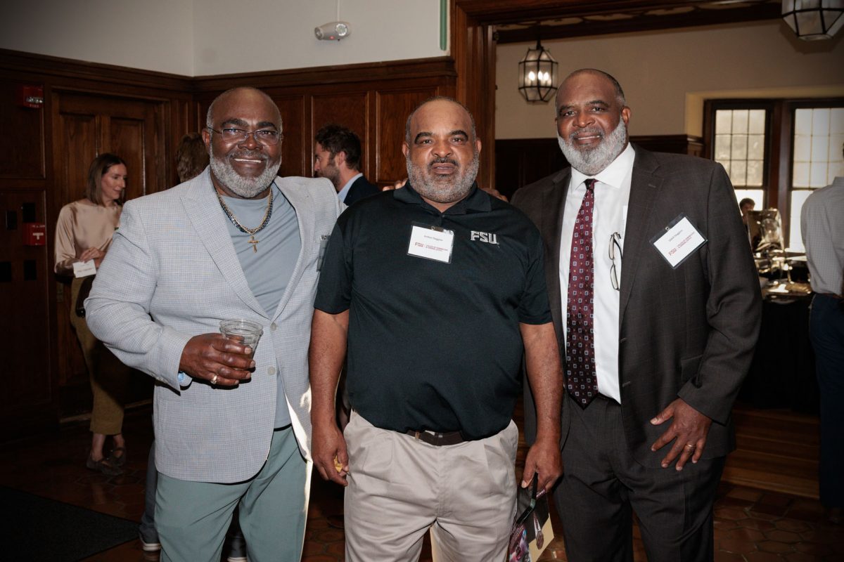 Odell Haggins, Jr. smiles while posing for a group photo with two men, one in a grey suit jacket and one in a black FSU polo shirt.
