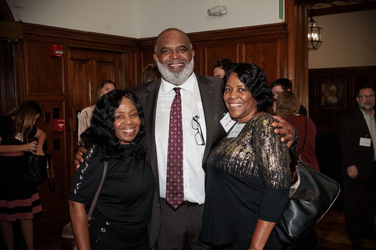 Odell Haggins, Jr. smiles with his arms around two women in a wood-paneled event space.