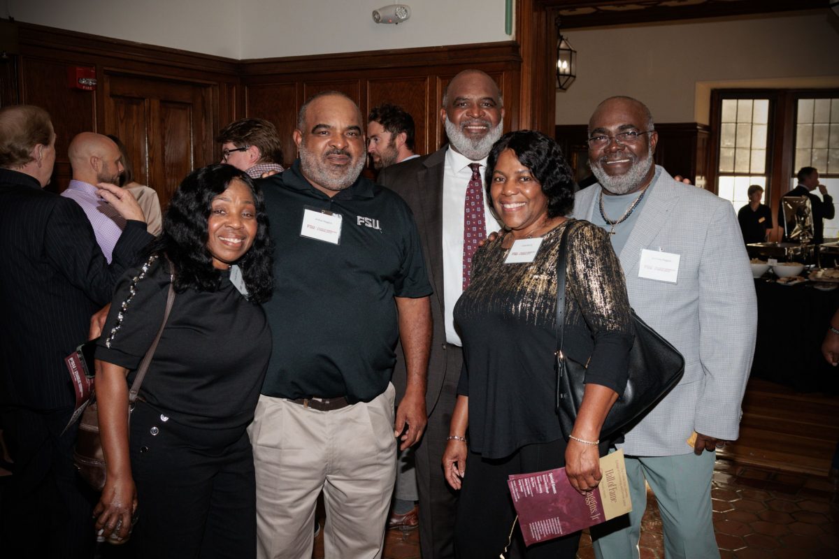 Odell Haggins, Jr. smiles while posing in a group group photo with four other event guests indoors in front of wood paneling.