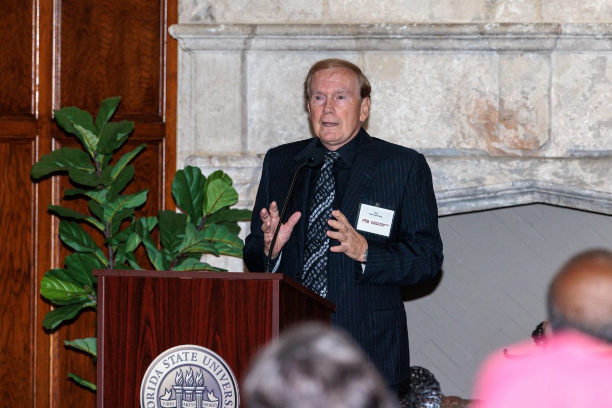 A man in a pinstripe suit speaks with raised hands at a Florida State University podium in front of a stone fireplace.