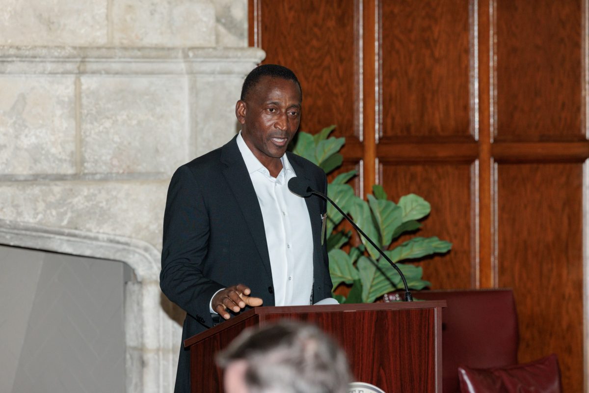 A man in a suit jacket and white dress shirt speaks at a podium with a microphone.