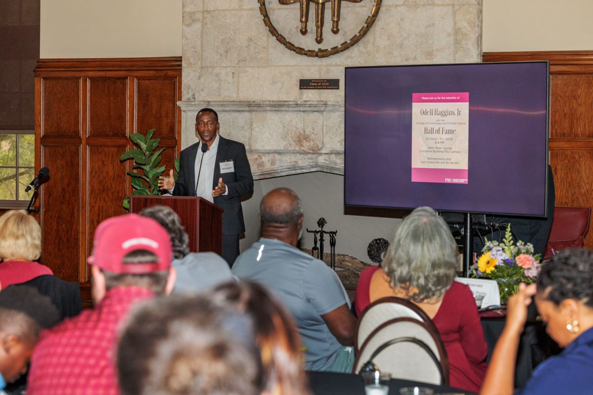 A man in a suit speaks at a podium to an audience in front of a presentation display for Odell Haggins Jr.'s Hall of Fame induction.