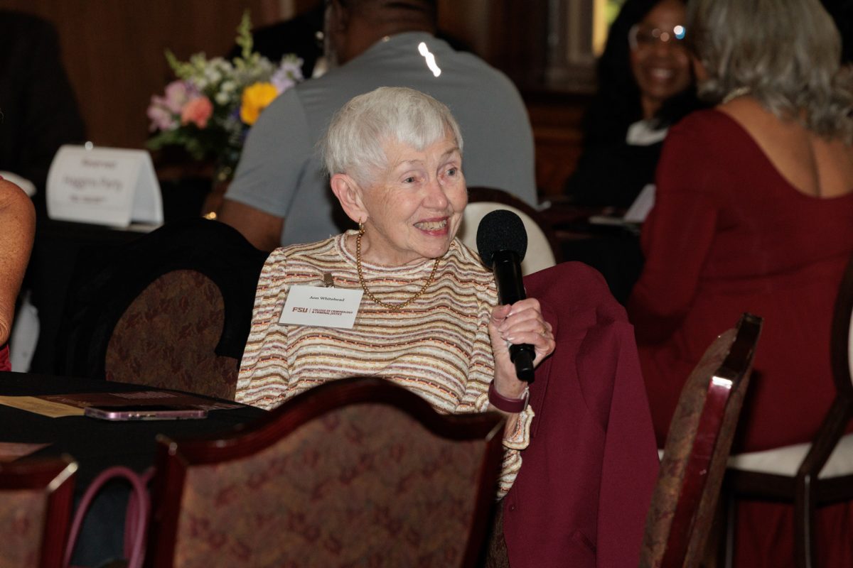 An older woman with short white hair smiles while holding a microphone at a table during an event presentation.
