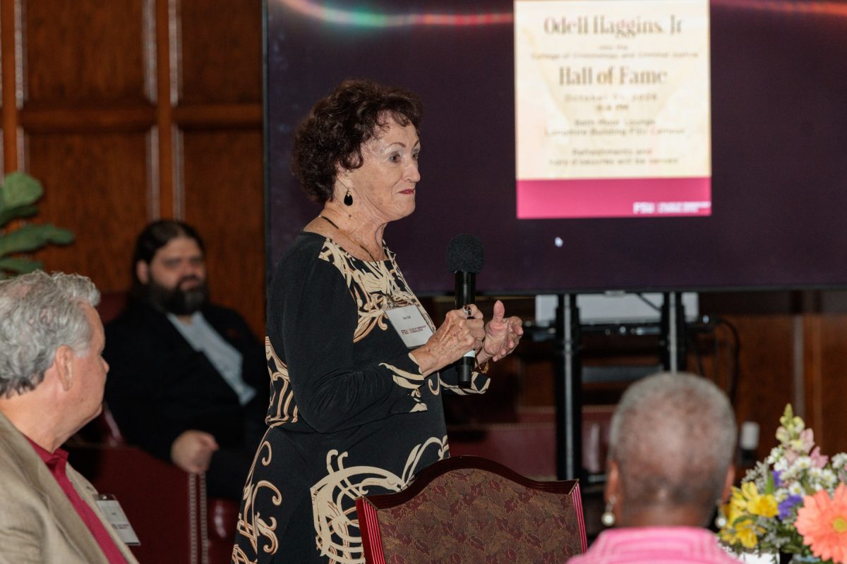A woman in a pattern dress speaks into a microphone in front of a presentation screen during a ceremony.