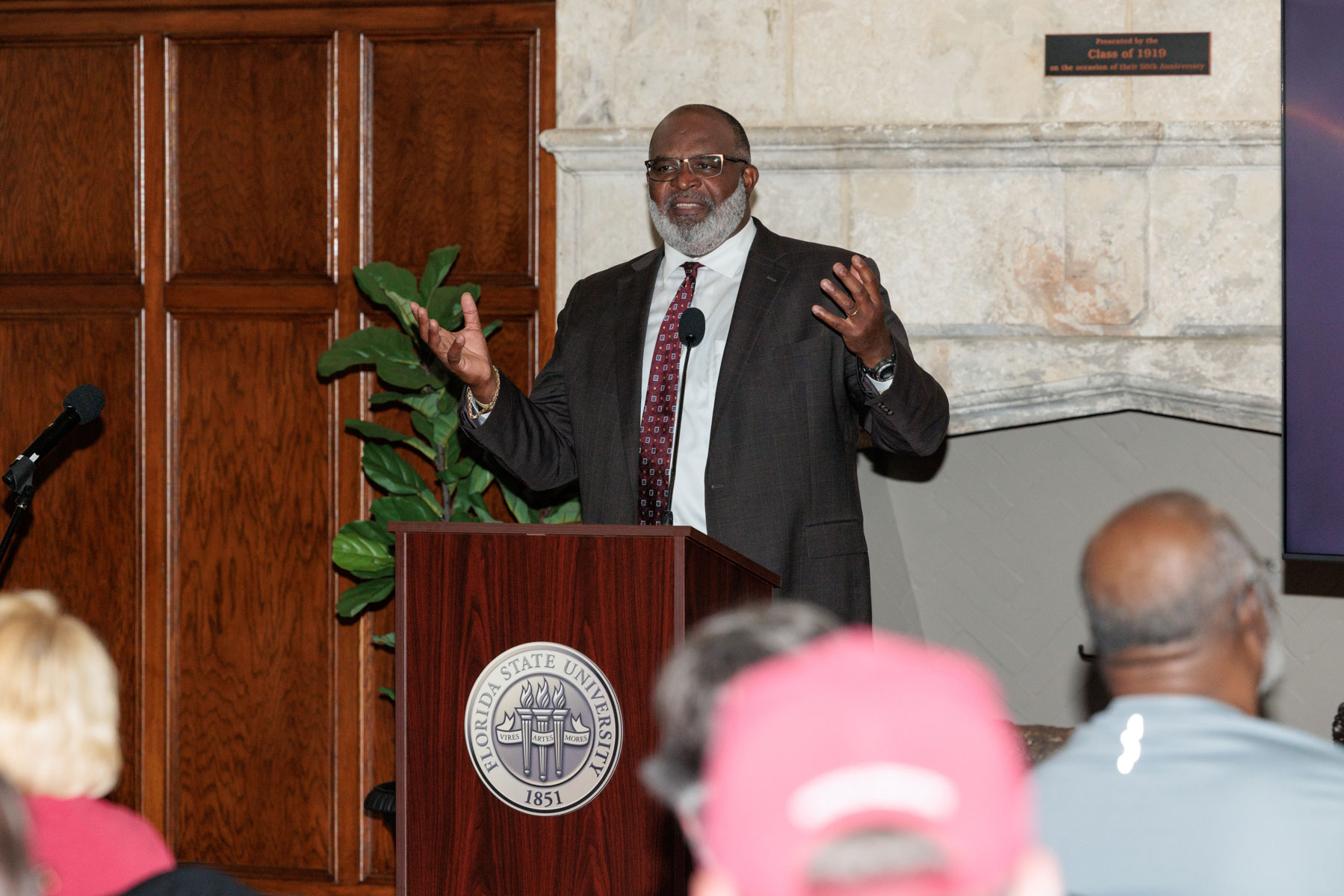 A man with a grey beard wearing a suit speaks at a Florida State University podium with his hands raised while addressing an audience.