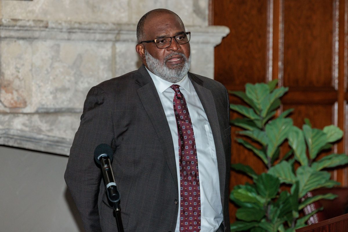 A man with a grey beard wearing glasses and a suit stands at a podium.