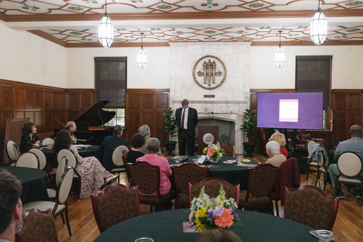 A man stands in front of a stone fireplace addressing guests seated at round tables in a decorated room.
