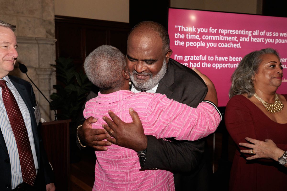 A smiling man in a suit warm-hugs a woman wearing a pink pattern jacket during an event presentation.