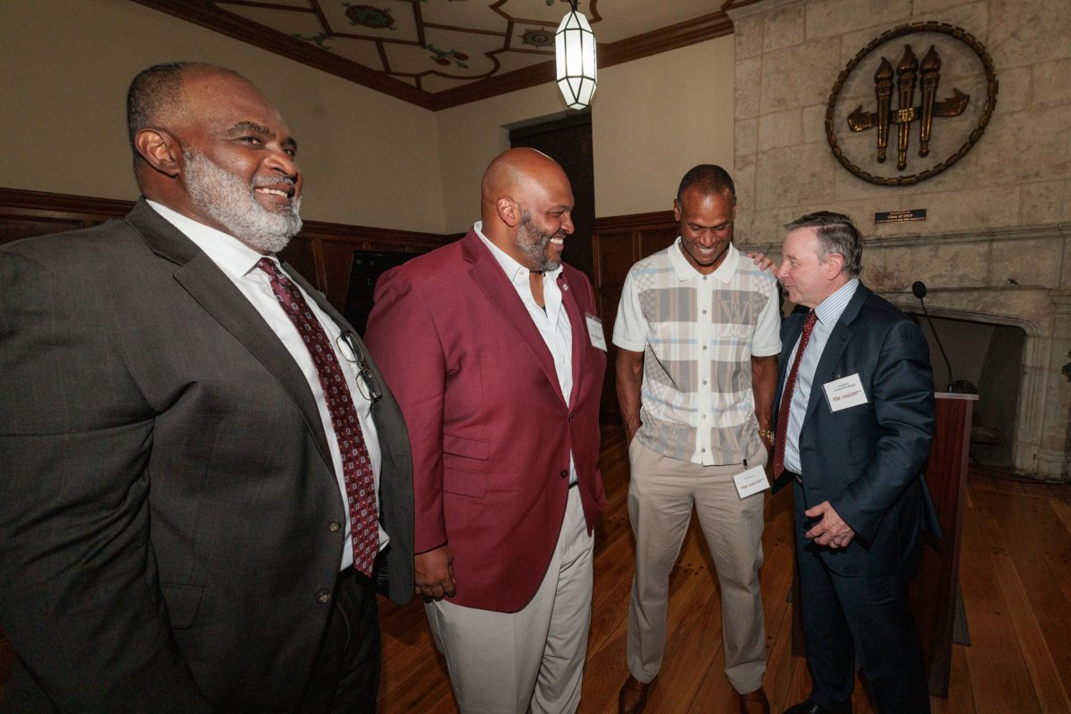Four men stand together in a room chatting and smiling, with a stone fireplace and seal visible in the background.