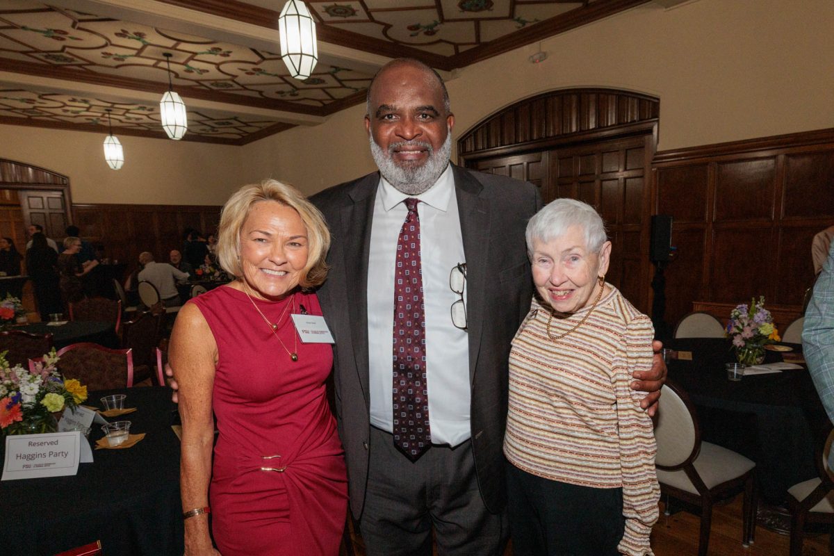 A smiling man in a suit poses between two women in an elegant event space with tables and chandeliers.