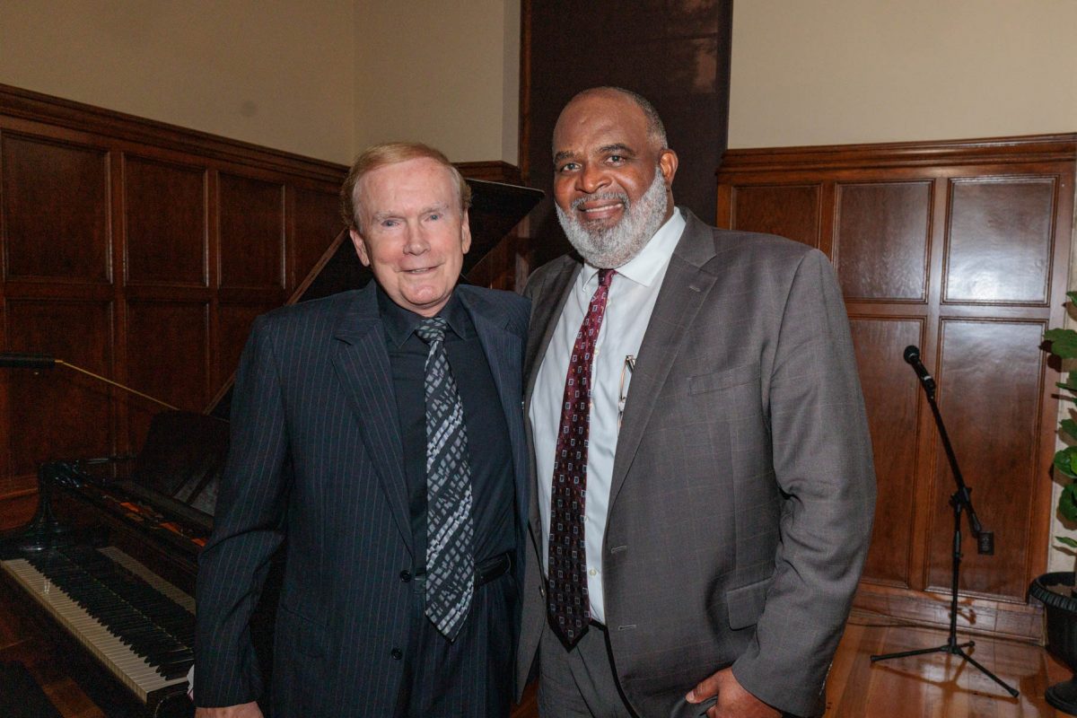 Two smiling men in suits pose together in front of a grand piano in a room with wood paneling.