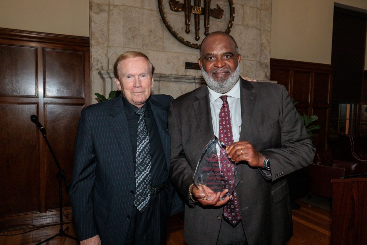 Two smiling men in suits stand next to each other, with the man on the right holding a glass award.