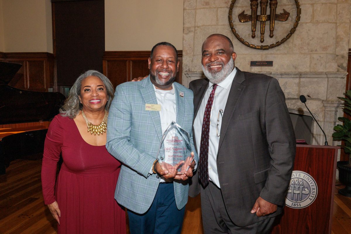 Three smiling people pose together as a man in a light blue plaid jacket holds up a clear crystal award next to Odell Haggins, Jr. in a dark suit.