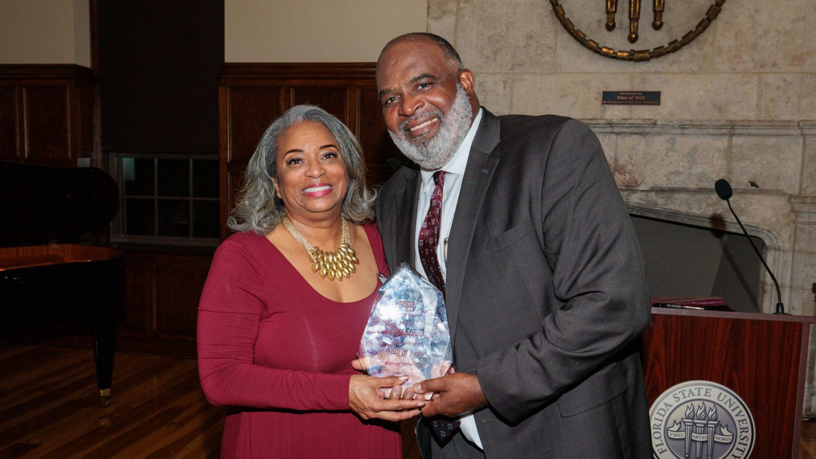 A smiling woman in a red dress and a man in a dark suit pose together while holding a crystal award in front of a Florida State University podium.