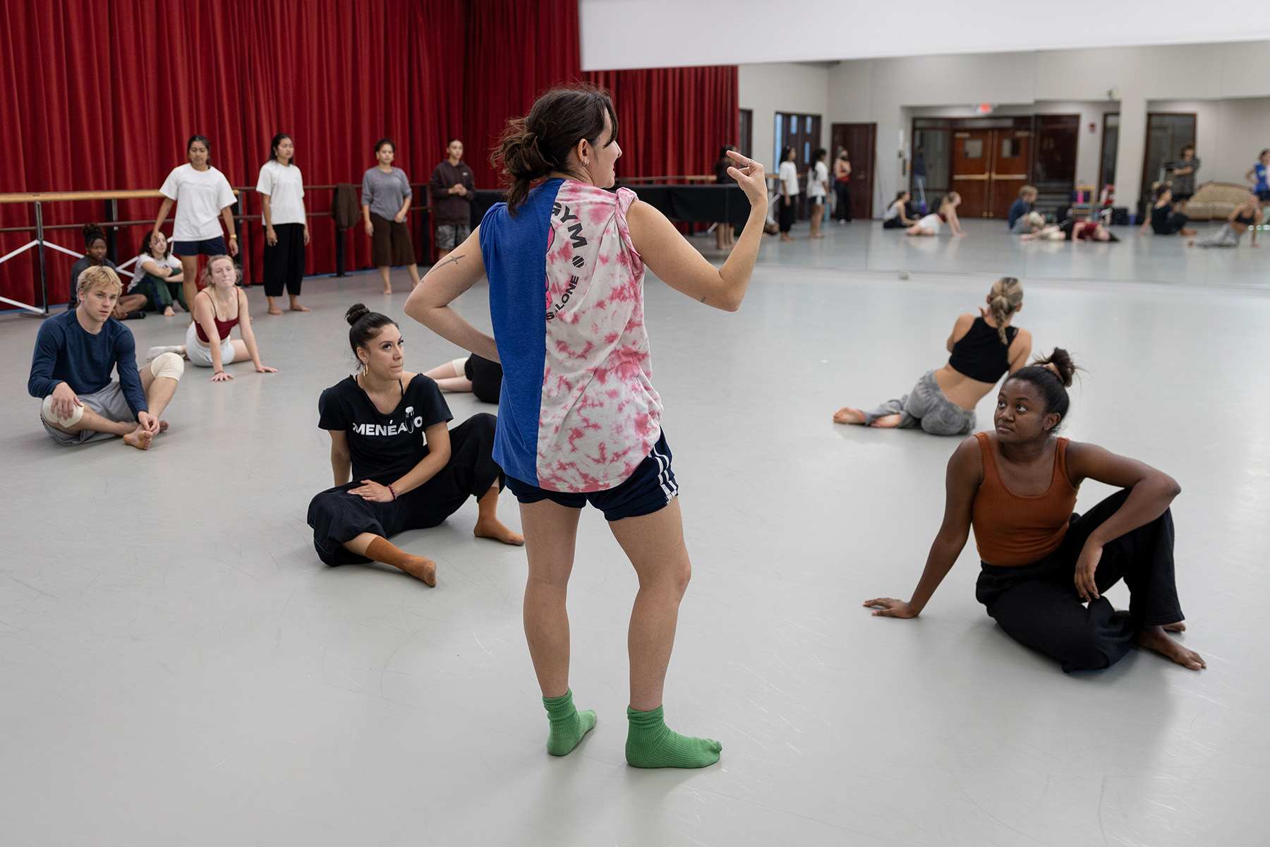 A wide shot of a dance studio where instructor Cecilia Benitez, viewed from behind, stands with one hand on her hip and gestures with her other hand toward the senior dance students seated on the studio floor listening.