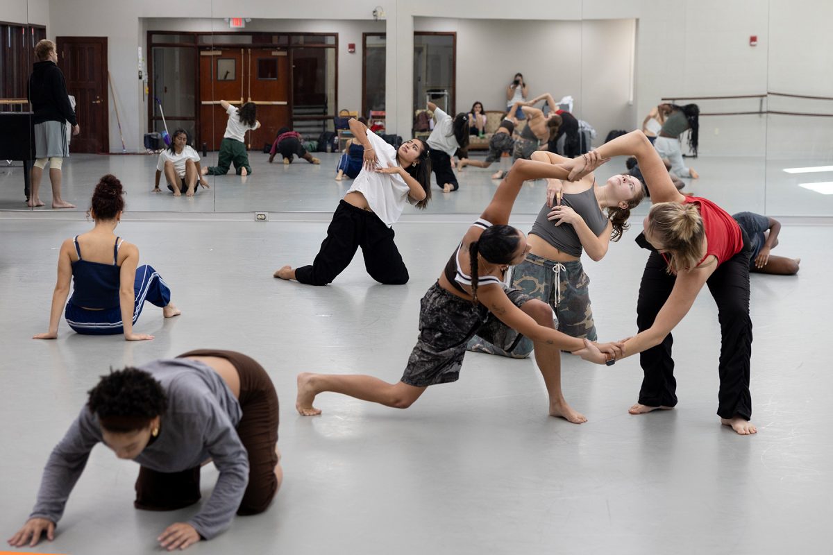 Numerous senior dancers are practicing contemporary dance moves in various group formations and individual poses in the dance studio, with a woman in the immediate foreground, facing down and toward the viewer, and in the center right, a complex trio is intertwined.