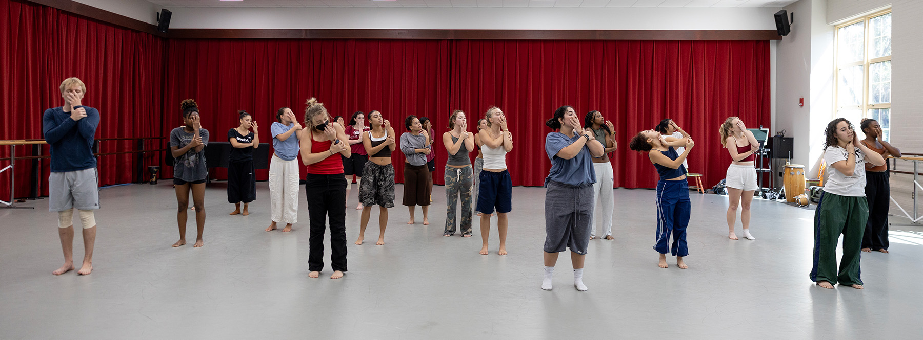 The senior cohort is scattered across the floor in a loose formation standing upright, and almost all of them are barefoot. Many of the dancers are in mid-movement, holding similar expressive poses that involve bringing their hands to or covering their faces.