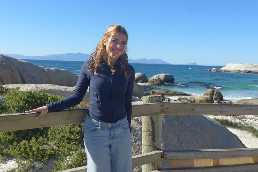 A young woman poses on Boulder Beach in South Africa with blue ocean water and mountains in the background.