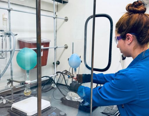A researcher wearing a blue lab coat and safety glasses adjusts chemical equipment fitted with balloons inside a fume hood in a laboratory.