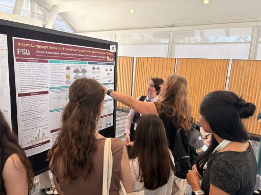 A presenter gestures towards a research poster titled "Infant Language Network Functional Connectivity Predicting Late Talking" while explaining the contents to a small group of attendees at an academic conference.