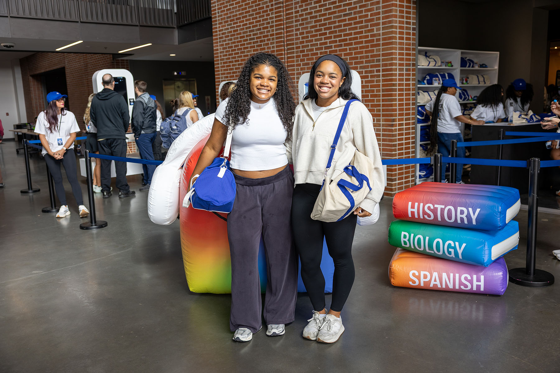 Two smiling young women pose together carrying shoulder bags in front of an event backdrop with giant inflatable books labeled "History," "Biology," and "Spanish."