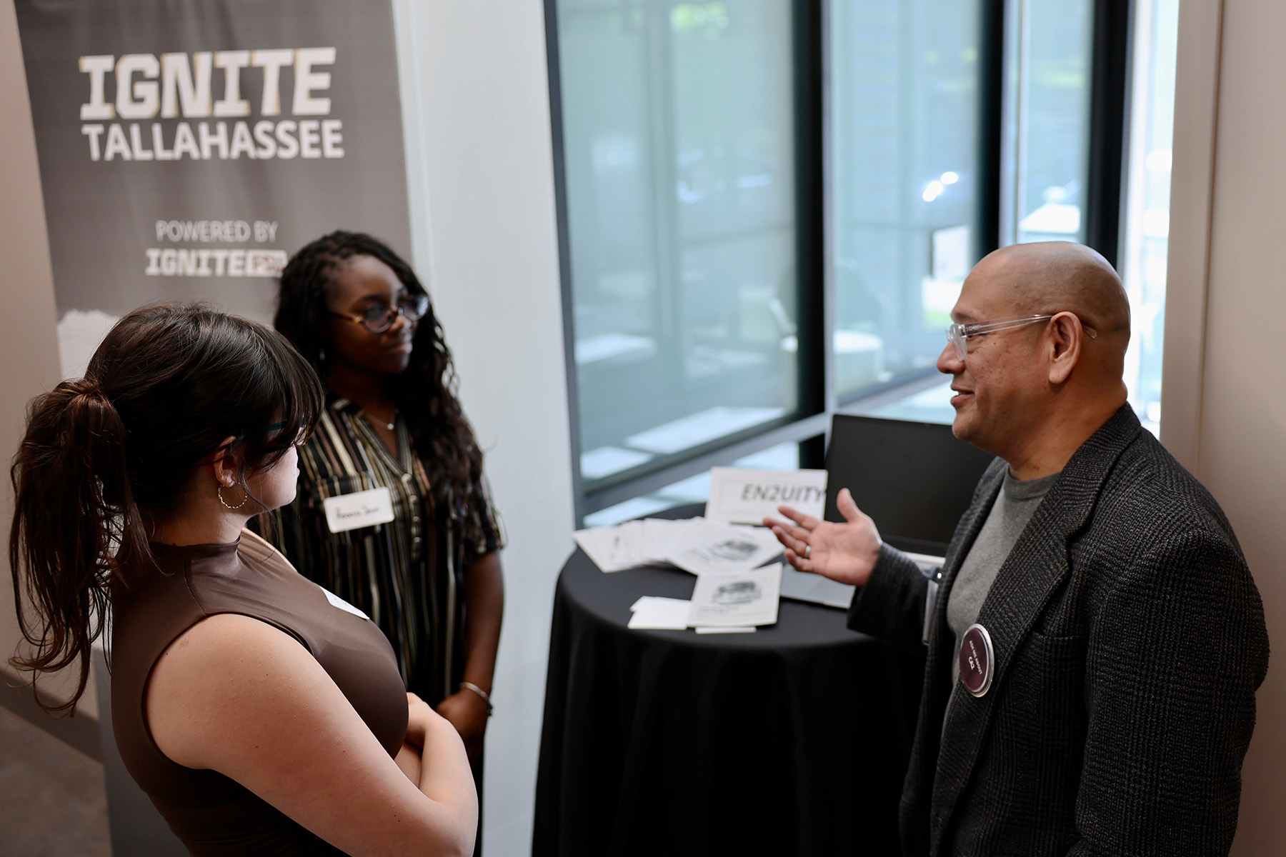 A man wearing glasses and a blazer speaks to two young women beside a table set up with informational materials near an "Ignite Tallahassee" banner.