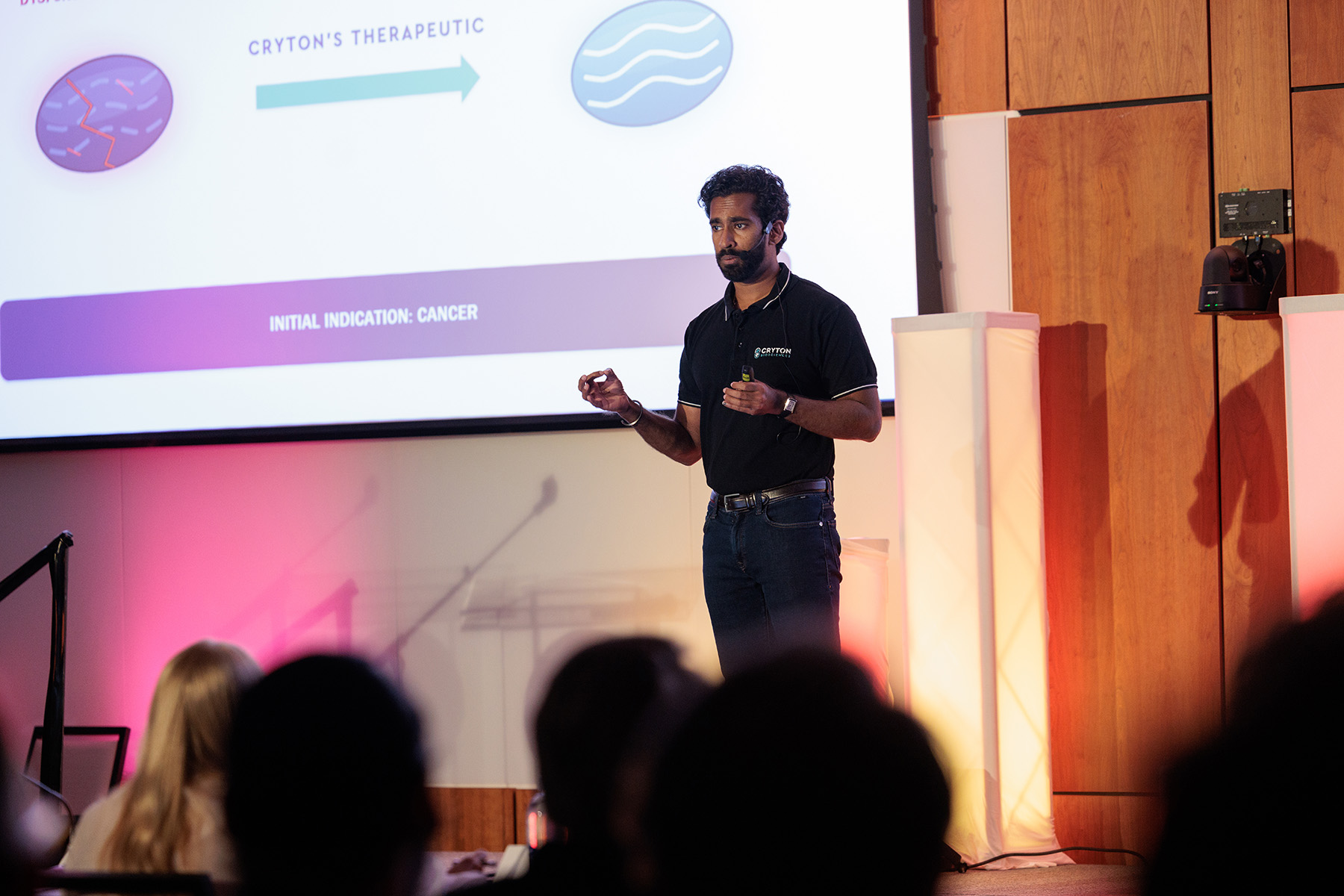 Anosh Gomes stands on stage presenting a powerpoint slide.