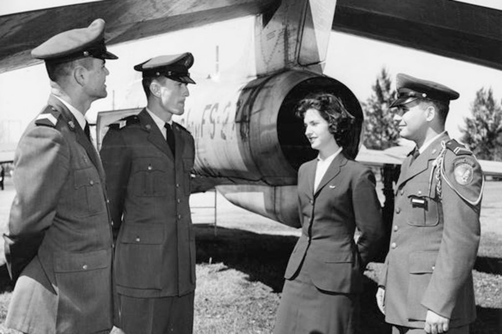 Black and white photograph of three men and one women standing in front of a plane in military uniforms.