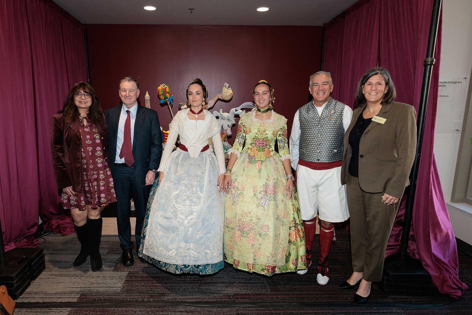 Six people stand in front of garnet curtains with a sculpture behind them.