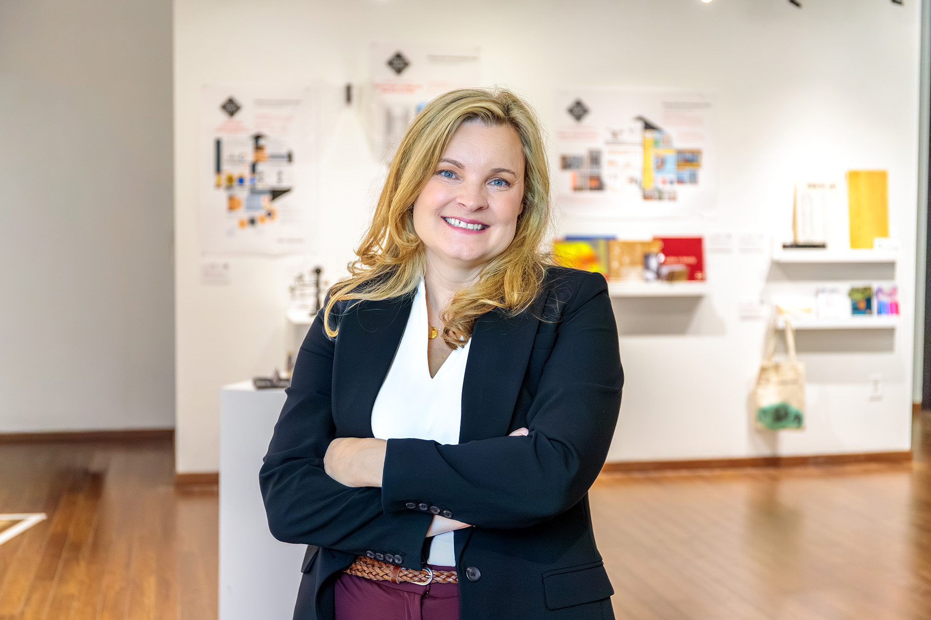 Amy Huber smiles with her arms crossed in a gallery space featuring design displays and exhibits.