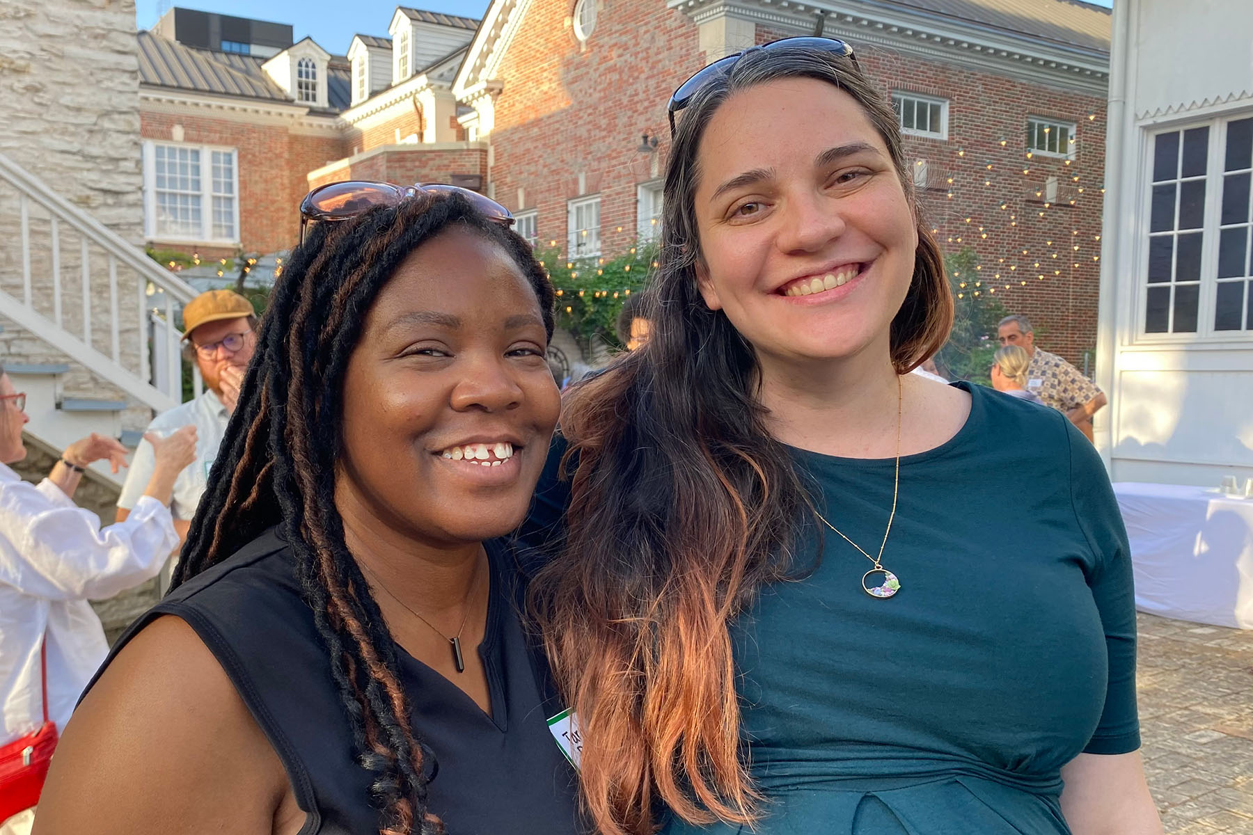 Two smiling women pose together outdoors during an event in front of a brick building.