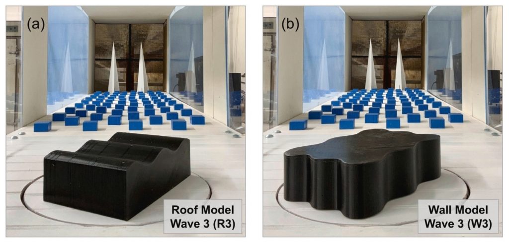 Side-by-side photographs of two black models mounted on a circular platform inside a wind tunnel. Panel (a) shows the angular Roof Model, labeled “Wave 3 (R3).” Panel (b) shows the rounded, wavy-edged Wall Model, labeled “Wave 3 (W3).” Rows of blue rectangular blocks and two white triangular structures are visible downstream behind each model.