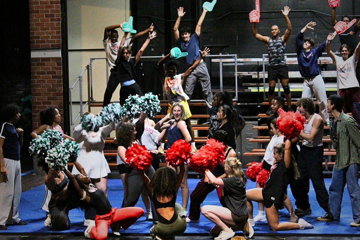 A large ensemble of performers rehearses a cheerleading number onstage, raising red and green pom-poms and foam fingers while others dance and pose on risers.