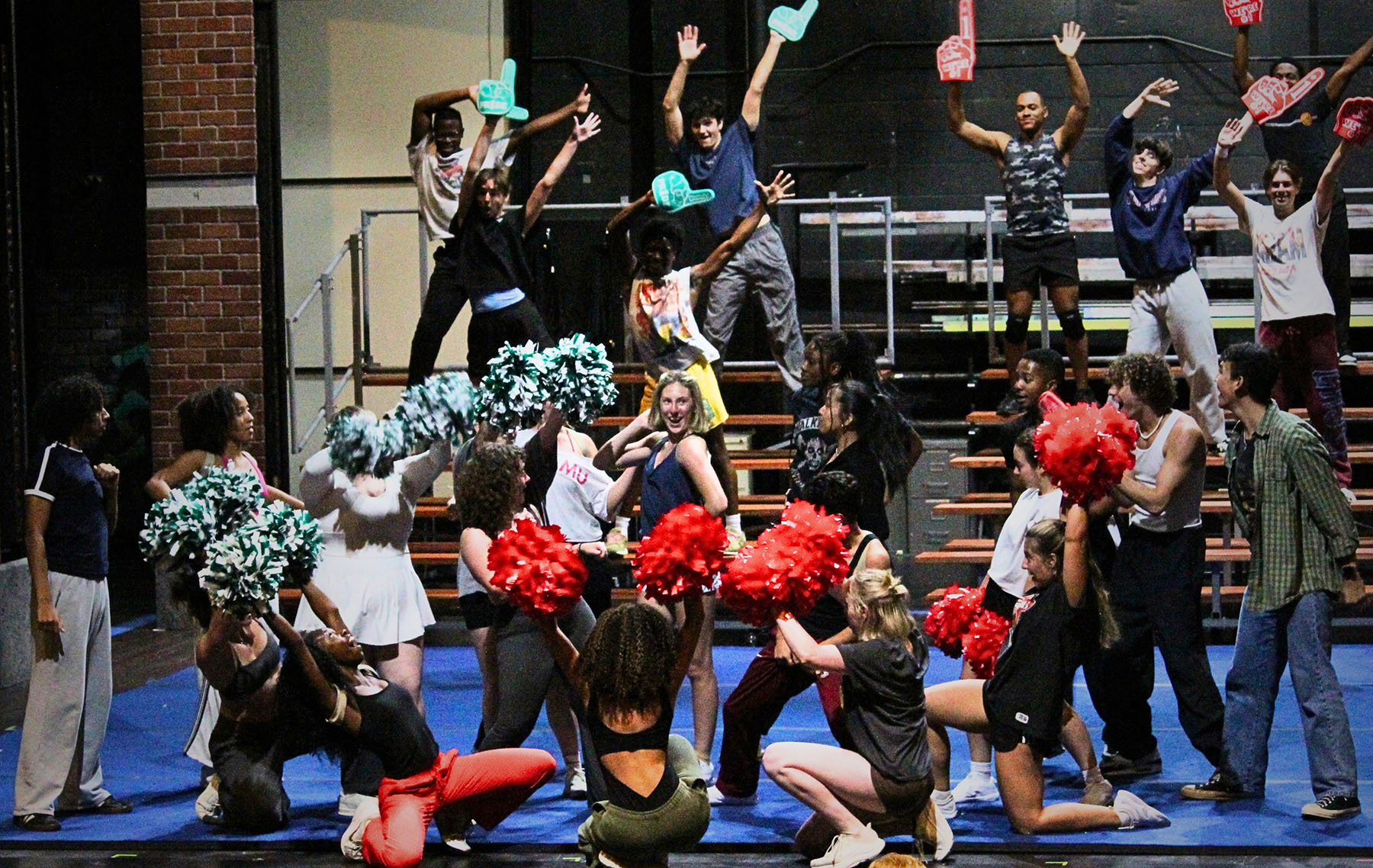 A large ensemble of performers rehearses a cheerleading number onstage, raising red and green pom-poms and foam fingers while others dance and pose on risers.