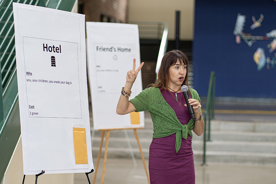 A presenter holding a microphone raises one finger while speaking beside oversized easel-mounted posters in a large indoor space. The nearest poster is labeled “Hotel” and includes brief information about who can use it and its cost.